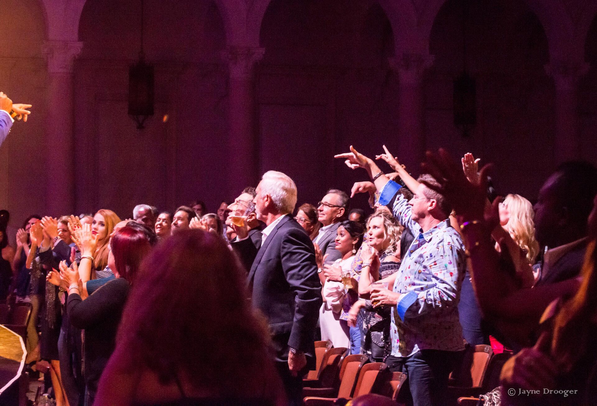 A theater audience stands, applauding and pointing toward the stage beneath arched walls during the concert