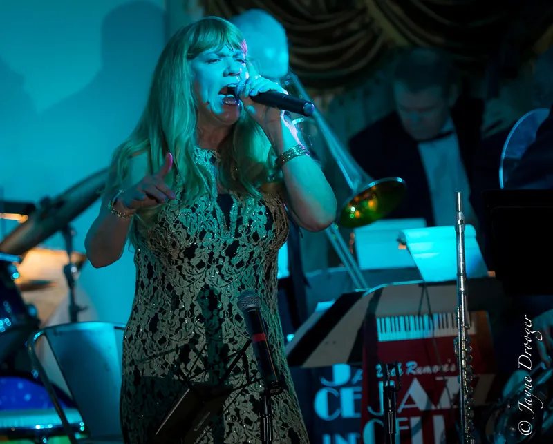 A vocalist with long blonde hair and a gold lace dress sings powerfully in front of the big band