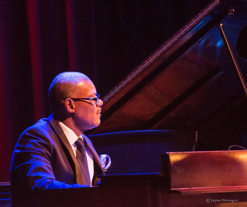 Shawn Brown, in glasses and a suit with a pocket square, plays a wooden organ in front of a grand piano